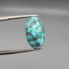 A 16.41-carat natural blue-green Turquoise (Feroza) from Kerman, Iran, featuring an oval cabochon cut with white quartz matrix and natural organic holes on both sides, held in jeweler's tweezers.