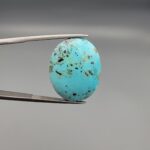 ​A 26.05-carat natural blue-green Turquoise (Feroza) from Kerman, Iran, featuring a symmetrical oval cabochon cut with a metallic golden pyrite matrix, held in jeweler's tweezers.