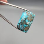 ​A 28.84-carat natural blue-green Turquoise (Feroza) from Kerman, Iran, featuring a unique rectangle cabochon cut with a shimmering golden pyrite matrix.