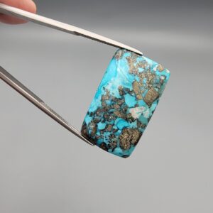 ​A 28.84-carat natural blue-green Turquoise (Feroza) from Kerman, Iran, featuring a unique rectangle cabochon cut with a shimmering golden pyrite matrix.