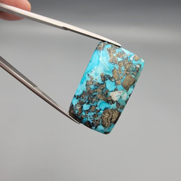 ​A 28.84-carat natural blue-green Turquoise (Feroza) from Kerman, Iran, featuring a unique rectangle cabochon cut with a shimmering golden pyrite matrix.