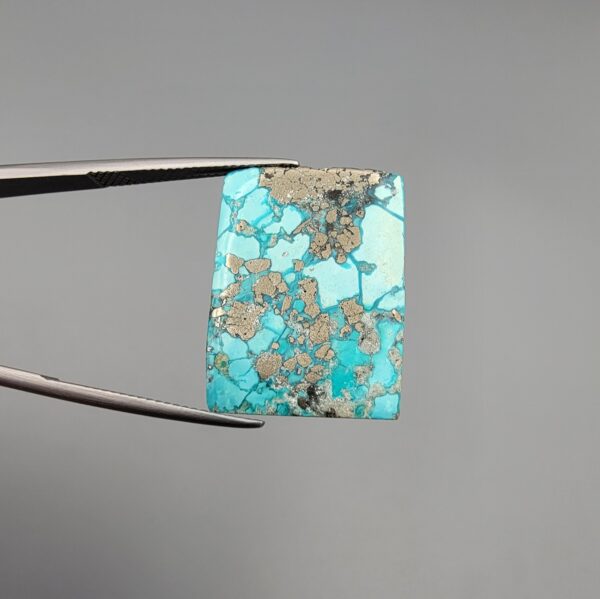 ​A 29.83-carat natural blue-green Turquoise (Feroza) from Kerman, Iran, featuring a unique rectangle cabochon cut with a shimmering golden pyrite matrix.