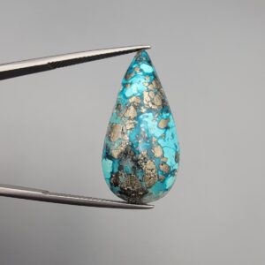 ​A 20.97-carat natural blue-green Turquoise (Feroza) from Kerman, Iran, featuring a pear-shaped cabochon cut with a shimmering golden pyrite matrix, held in jeweler's tweezers.