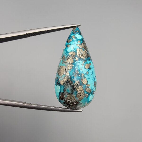 ​A 20.97-carat natural blue-green Turquoise (Feroza) from Kerman, Iran, featuring a pear-shaped cabochon cut with a shimmering golden pyrite matrix, held in jeweler's tweezers.