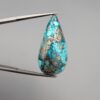 ​A 20.97-carat natural blue-green Turquoise (Feroza) from Kerman, Iran, featuring a pear-shaped cabochon cut with a shimmering golden pyrite matrix, held in jeweler's tweezers.