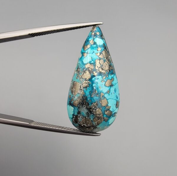 ​A 20.97-carat natural blue-green Turquoise (Feroza) from Kerman, Iran, featuring a pear-shaped cabochon cut with a shimmering golden pyrite matrix, held in jeweler's tweezers.