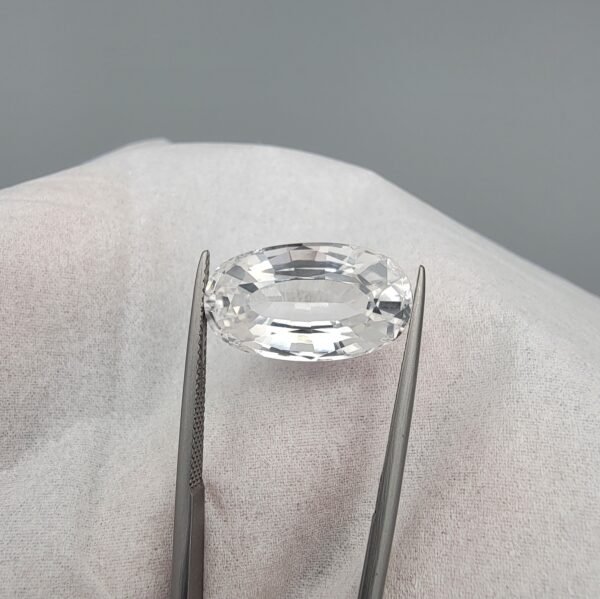 ​A large 14.78-carat natural Kunzite gemstone from Pakistan, featuring an oval faceted cut, eye-clean clarity, and a light pink to white color-shift effect, held in jeweler's tweezers.