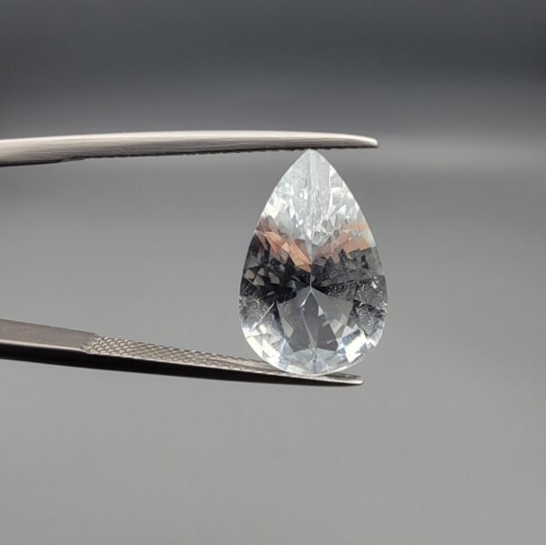​A large 5.35-carat natural Shigar Valley Aquamarine (Beruj) gemstone with a pear-shaped faceted cut, displaying an icy-blue color and organic mountain inclusions, held in jeweler's tweezers.