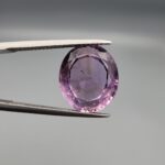 ​A 7.89-carat natural Brazilian Amethyst (Katela) gemstone with an oval faceted cut, featuring a deep purple color and high clarity, held in jeweler's tweezers.