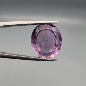 ​A 7.89-carat natural Brazilian Amethyst (Katela) gemstone with an oval faceted cut, featuring a deep purple color and high clarity, held in jeweler's tweezers.