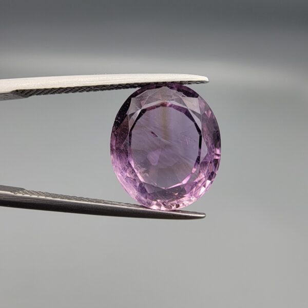 ​A 7.89-carat natural Brazilian Amethyst (Katela) gemstone with an oval faceted cut, featuring a deep purple color and high clarity, held in jeweler's tweezers.