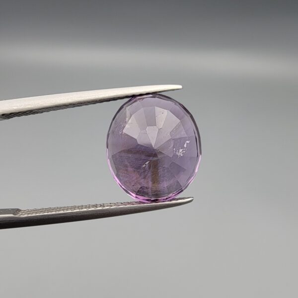​A 7.89-carat natural Brazilian Amethyst (Katela) gemstone with an oval faceted cut, featuring a deep purple color and high clarity, held in jeweler's tweezers.