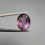 ​A 6.89-carat natural Brazilian Amethyst (Katela) gemstone with a professional oval faceted cut, displaying a deep purple hue and high transparency, held in jeweler's tweezers.