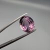 ​A 6.89-carat natural Brazilian Amethyst (Katela) gemstone with a professional oval faceted cut, displaying a deep purple hue and high transparency, held in jeweler's tweezers.