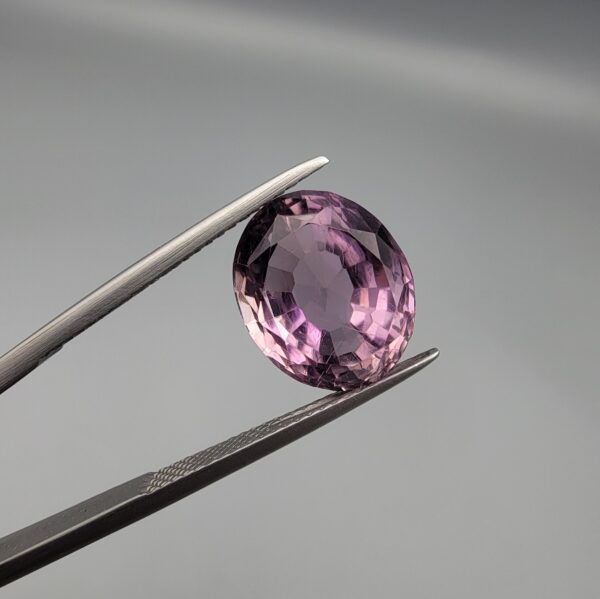 ​A 6.89-carat natural Brazilian Amethyst (Katela) gemstone with a professional oval faceted cut, displaying a deep purple hue and high transparency, held in jeweler's tweezers.