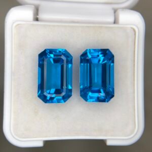 ​A matching pair of natural Swiss Blue Topaz gemstones featuring a sophisticated emerald cut and a total combined weight of 16.87 carats, displayed in a white jeweler’s box.