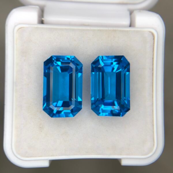 ​A matching pair of natural Swiss Blue Topaz gemstones featuring a sophisticated emerald cut and a total combined weight of 16.87 carats, displayed in a white jeweler’s box.