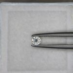 A 0.13-carat natural white diamond (Heera) with a round brilliant cut, displaying sharp faceting and intense sparkle, held securely in jeweler's tweezers.