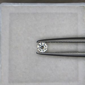 A 0.13-carat natural white diamond (Heera) with a round brilliant cut, displaying sharp faceting and intense sparkle, held securely in jeweler's tweezers.