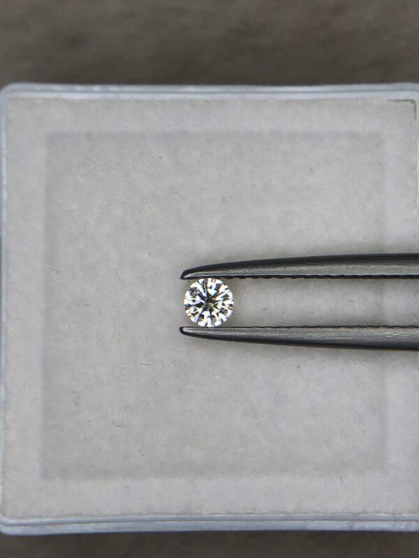 A 0.13-carat natural white diamond (Heera) with a round brilliant cut, displaying sharp faceting and intense sparkle, held securely in jeweler's tweezers.