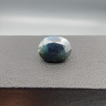 A 5.10-carat natural sapphire from Kanchanaburi, Thailand, featuring a unique blue-green color blend and an oval faceted cut, displayed in a white jeweler's box.