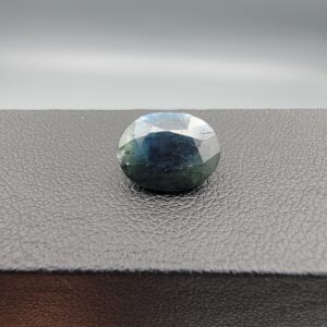 A 5.10-carat natural sapphire from Kanchanaburi, Thailand, featuring a unique blue-green color blend and an oval faceted cut, displayed in a white jeweler's box.