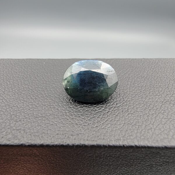 A 5.10-carat natural sapphire from Kanchanaburi, Thailand, featuring a unique blue-green color blend and an oval faceted cut, displayed in a white jeweler's box.