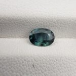 A 0.68-carat natural teal blue sapphire from Kanchanaburi, Thailand, featuring an oval faceted cut and a unique blue-green hue, displayed in a professional jeweler's box