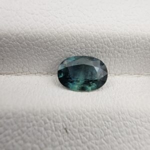 A 0.68-carat natural teal blue sapphire from Kanchanaburi, Thailand, featuring an oval faceted cut and a unique blue-green hue, displayed in a professional jeweler's box