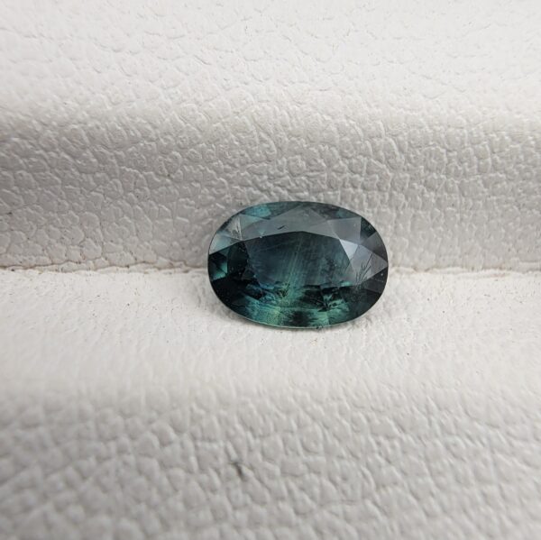 A 0.68-carat natural teal blue sapphire from Kanchanaburi, Thailand, featuring an oval faceted cut and a unique blue-green hue, displayed in a professional jeweler's box