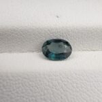 A 0.91-carat natural teal blue sapphire from Kanchanaburi, Thailand, featuring a classic oval faceted cut and a unique blue-green saturation, displayed in a professional white jeweler's box.