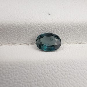 A 0.91-carat natural teal blue sapphire from Kanchanaburi, Thailand, featuring a classic oval faceted cut and a unique blue-green saturation, displayed in a professional white jeweler's box.