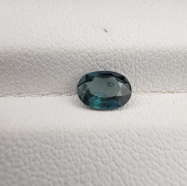 A 0.91-carat natural teal blue sapphire from Kanchanaburi, Thailand, featuring a classic oval faceted cut and a unique blue-green saturation, displayed in a professional white jeweler's box.