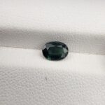 A 0.96-carat natural teal blue sapphire from Kanchanaburi, Thailand, featuring a classic oval faceted cut and a saturated blue-green color, presented in a professional white jeweler's box.