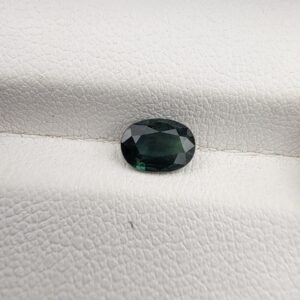 A 0.96-carat natural teal blue sapphire from Kanchanaburi, Thailand, featuring a classic oval faceted cut and a saturated blue-green color, presented in a professional white jeweler's box.