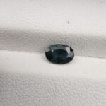 A 0.87-carat natural teal blue sapphire from Kanchanaburi, Thailand, featuring a classic oval faceted cut and a saturated blue-green color, presented in a professional white jeweler's box.