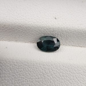 A 0.87-carat natural teal blue sapphire from Kanchanaburi, Thailand, featuring a classic oval faceted cut and a saturated blue-green color, presented in a professional white jeweler's box.