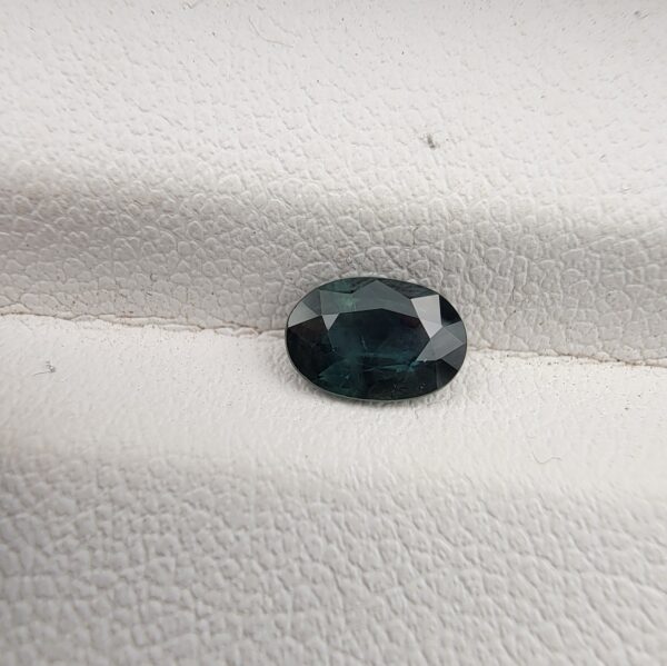 A 0.87-carat natural teal blue sapphire from Kanchanaburi, Thailand, featuring a classic oval faceted cut and a saturated blue-green color, presented in a professional white jeweler's box.