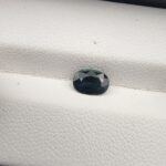 A 0.96-carat natural teal blue sapphire from Kanchanaburi, Thailand, featuring a masterfully faceted oval cut and deep blue-green saturation, presented in a professional white jeweler's box.