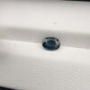 A 0.85-carat natural teal blue sapphire from Kanchanaburi, Thailand, with a classic oval faceted cut and saturated blue-green color, presented in a professional white jeweler's box.