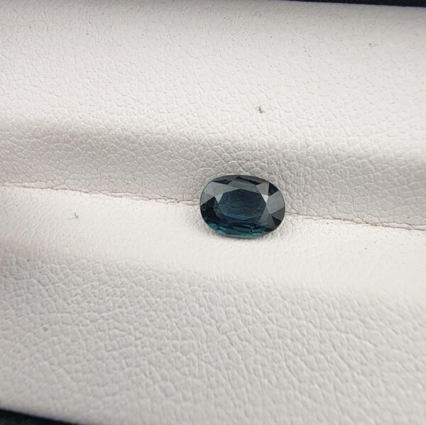 A 0.85-carat natural teal blue sapphire from Kanchanaburi, Thailand, with a classic oval faceted cut and saturated blue-green color, presented in a professional white jeweler's box.