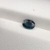 A 0.85-carat natural teal blue sapphire from Kanchanaburi, Thailand, with a classic oval faceted cut and saturated blue-green color, presented in a professional white jeweler's box.