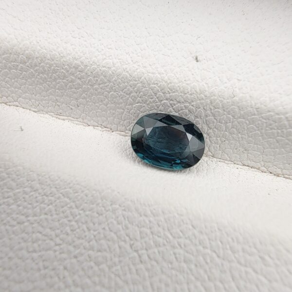 A 0.85-carat natural teal blue sapphire from Kanchanaburi, Thailand, with a classic oval faceted cut and saturated blue-green color, presented in a professional white jeweler's box.