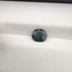 A 0.85-carat natural teal blue sapphire from Kanchanaburi, Thailand, with a classic oval faceted cut and saturated blue-green color, presented in a professional white jeweler's box.