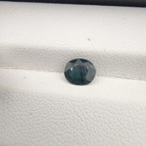 A 0.85-carat natural teal blue sapphire from Kanchanaburi, Thailand, with a classic oval faceted cut and saturated blue-green color, presented in a professional white jeweler's box.