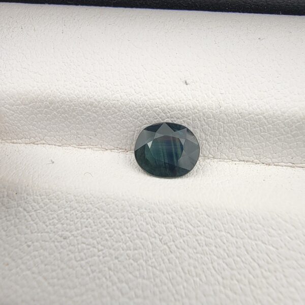 A 0.85-carat natural teal blue sapphire from Kanchanaburi, Thailand, with a classic oval faceted cut and saturated blue-green color, presented in a professional white jeweler's box.