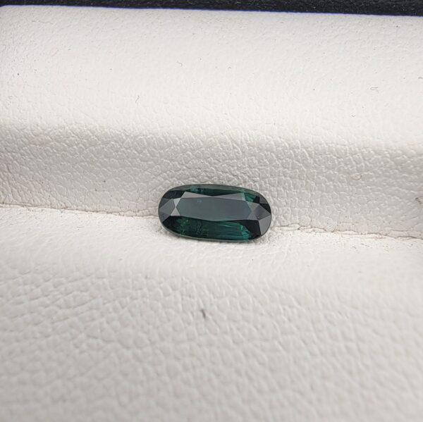 ​A 0.89-carat natural teal blue sapphire from Kanchanaburi, Thailand, featuring a classic oval faceted cut and saturated blue-green color, presented in a professional white jeweler's box.