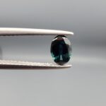 A 0.61-carat natural teal blue sapphire from Kanchanaburi, Thailand, featuring a classic oval faceted cut and a saturated blue-green color, presented in a professional white jeweler's box.