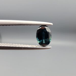 A 0.61-carat natural teal blue sapphire from Kanchanaburi, Thailand, featuring a classic oval faceted cut and a saturated blue-green color, presented in a professional white jeweler's box.