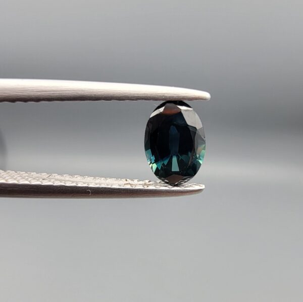A 0.61-carat natural teal blue sapphire from Kanchanaburi, Thailand, featuring a classic oval faceted cut and a saturated blue-green color, presented in a professional white jeweler's box.